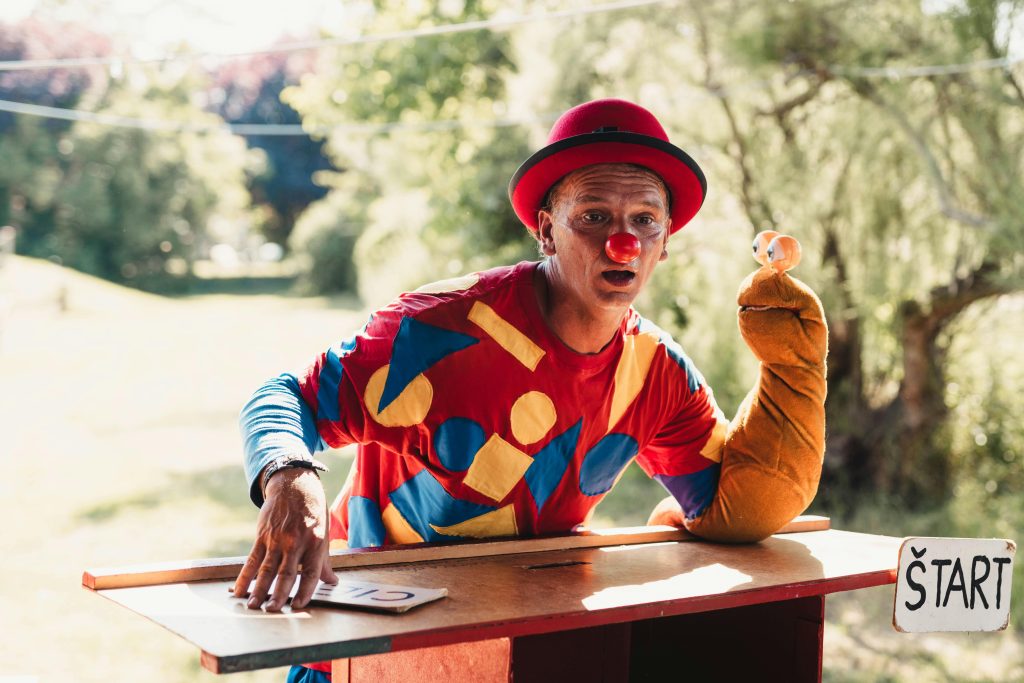 A clown wearing a colorful costume entertains with a puppet outdoors on a sunny day.